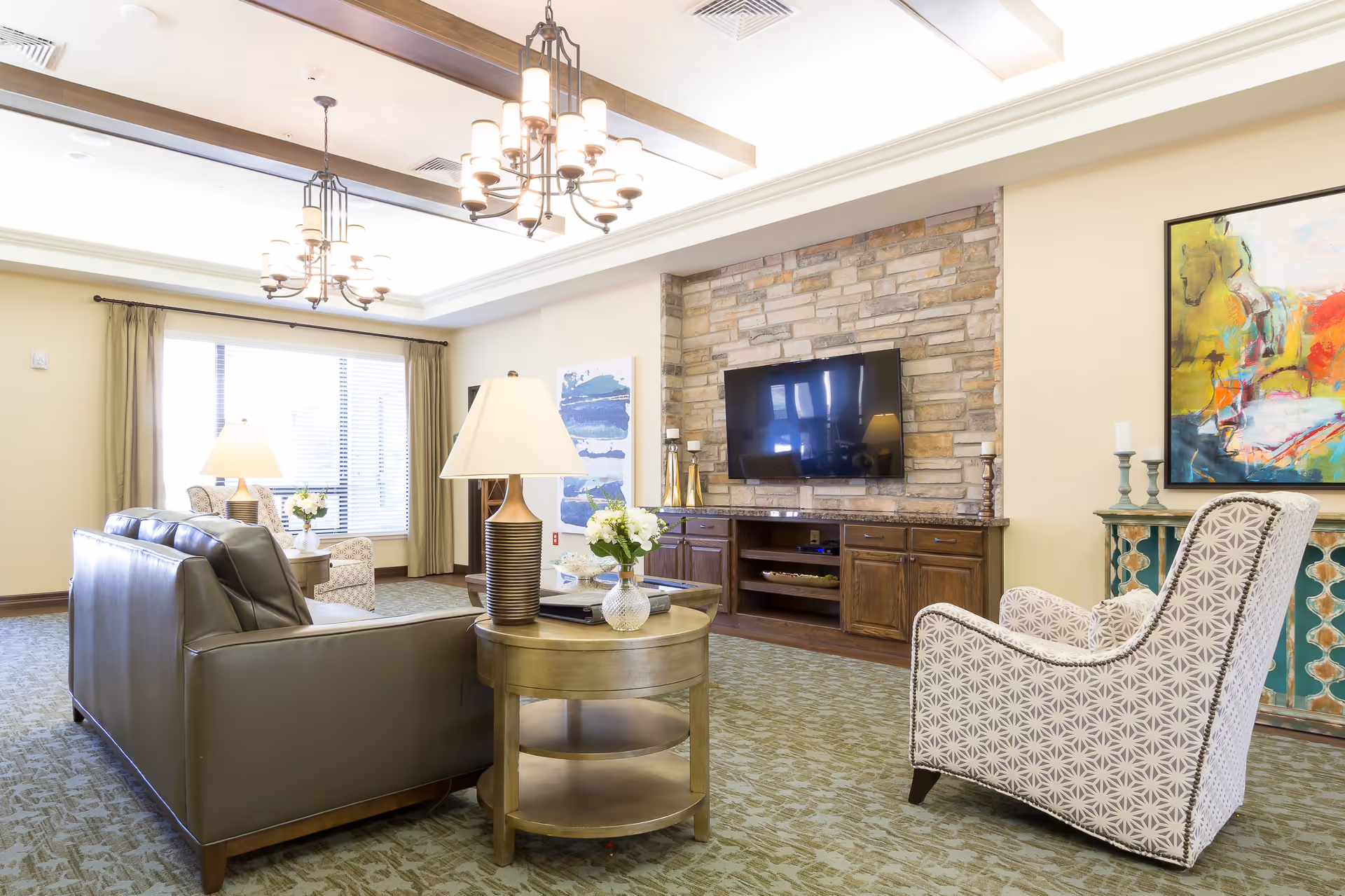 A bright and spacious senior living facility lounge area with a leather sofa, patterned armchair, round wooden side table with a lamp and flowers, a stone accent wall with a mounted flat-screen TV, and colorful artwork on the walls. Large windows with curtains allow natural light to fill the room.