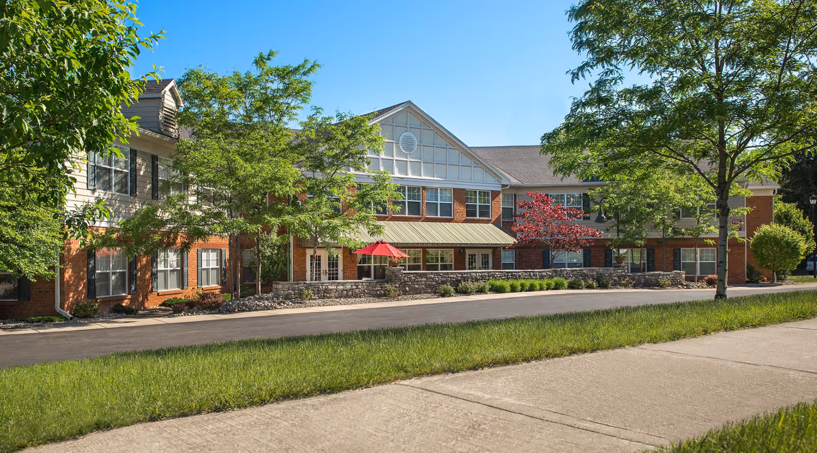 Exterior view of a two-story senior living facility building with brick and siding facade, surrounded by green trees and landscaping under a clear blue sky. A sidewalk and a paved driveway are visible in the foreground.