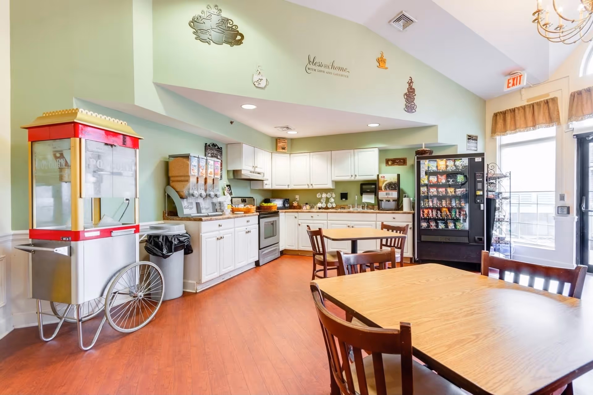 A bright and clean dining area with wooden tables and chairs, a popcorn machine on wheels, a snack vending machine, and a kitchen area with white cabinets, a stove, and various beverage dispensers. The walls are light green with decorative coffee-themed wall art and a sign that reads 'Bless this home with love and laughter'. There is a large window and a glass door letting in natural light.