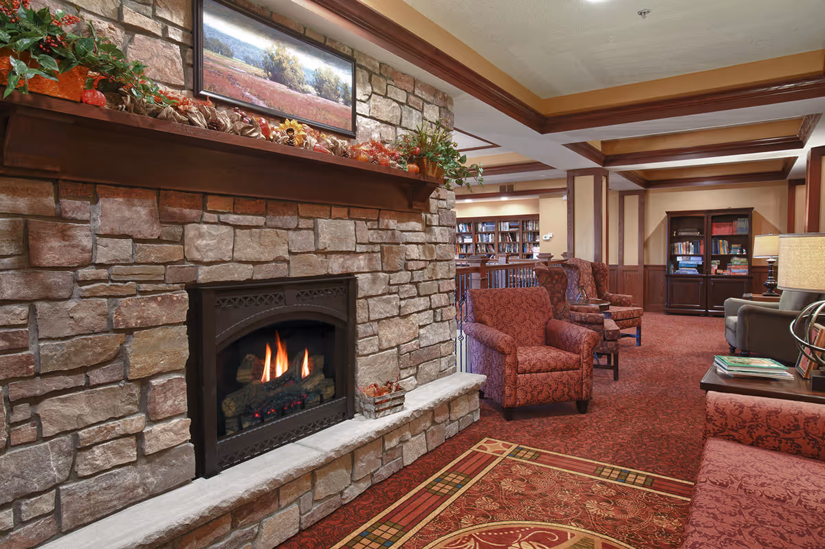 Cozy senior living lounge with a stone fireplace, patterned armchairs, bookshelves, and warm wood trim.