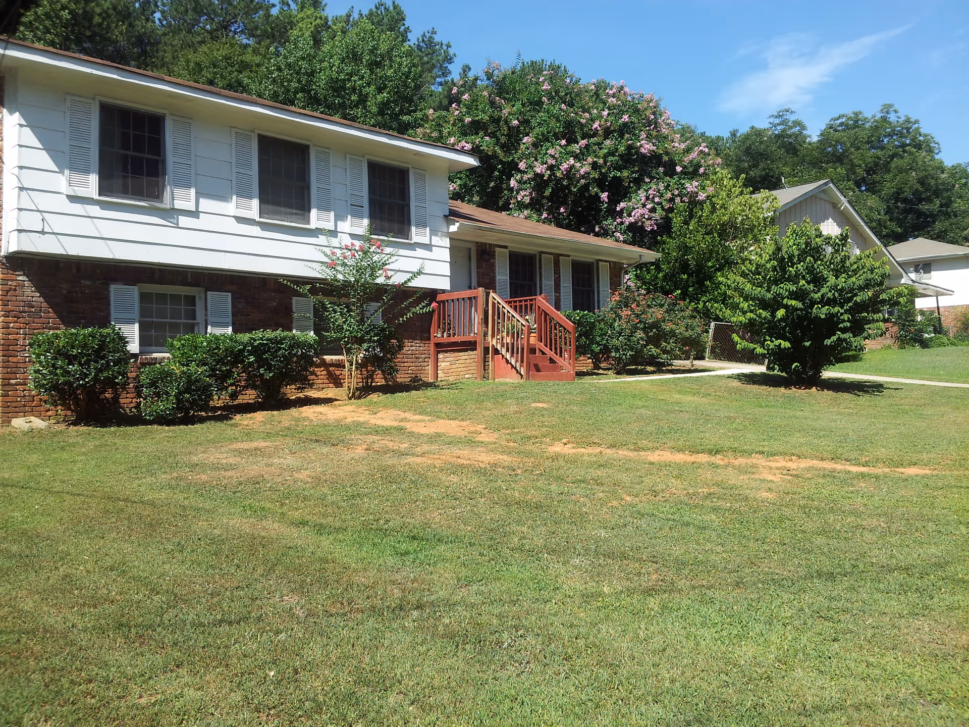 Exterior view of a residential building with a brick lower level and white siding upper level. The building has several windows with white shutters and a small porch with red wooden stairs. The lawn in front is green with some patches of dry grass, and there are bushes and trees around the building under a clear blue sky.