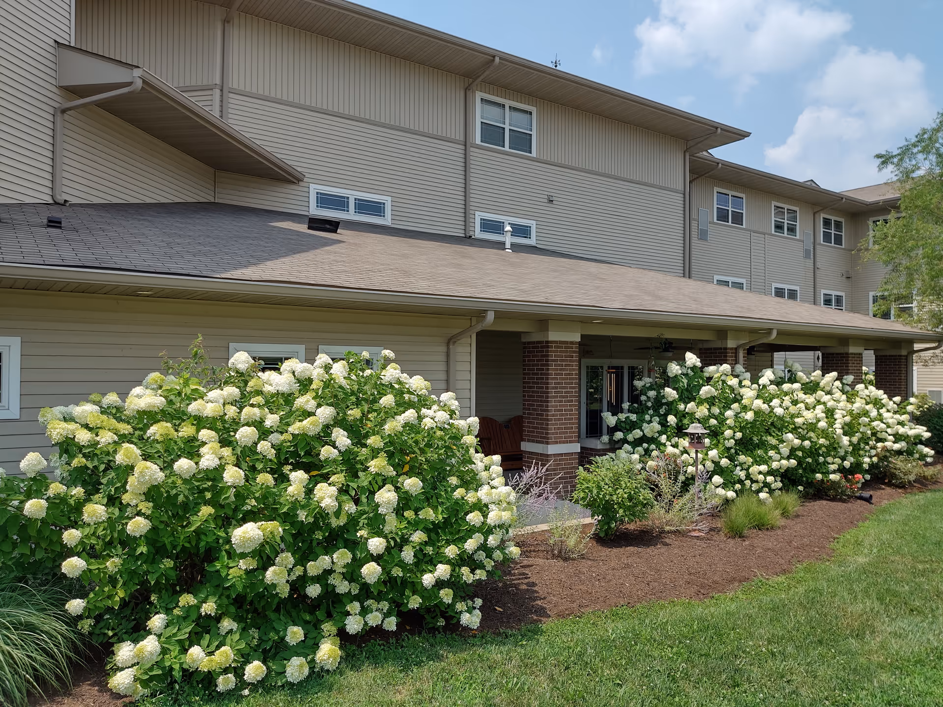 Exterior view of a senior living facility building with beige siding and a brown roof. The building has multiple windows and a covered porch area supported by brick columns. In front of the building, there are large bushes with white flowers and a well-maintained lawn under a partly cloudy sky.