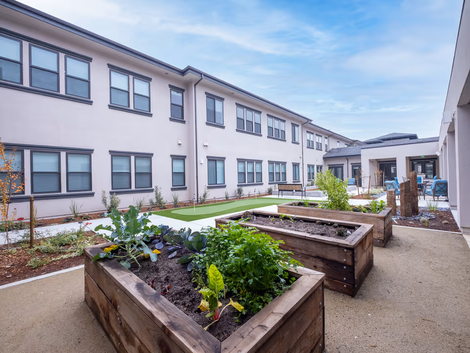 Courtyard of a two-story senior living facility featuring raised wooden garden beds, seating areas, and a small putting green.