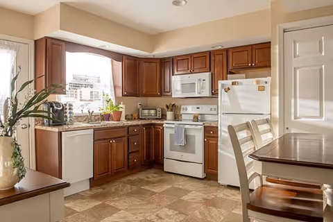 A bright kitchen with wooden cabinets, a white refrigerator, stove, microwave, and dishwasher. There is a window above the sink with potted plants on the countertop. A small dining table with chairs is visible on the right side.