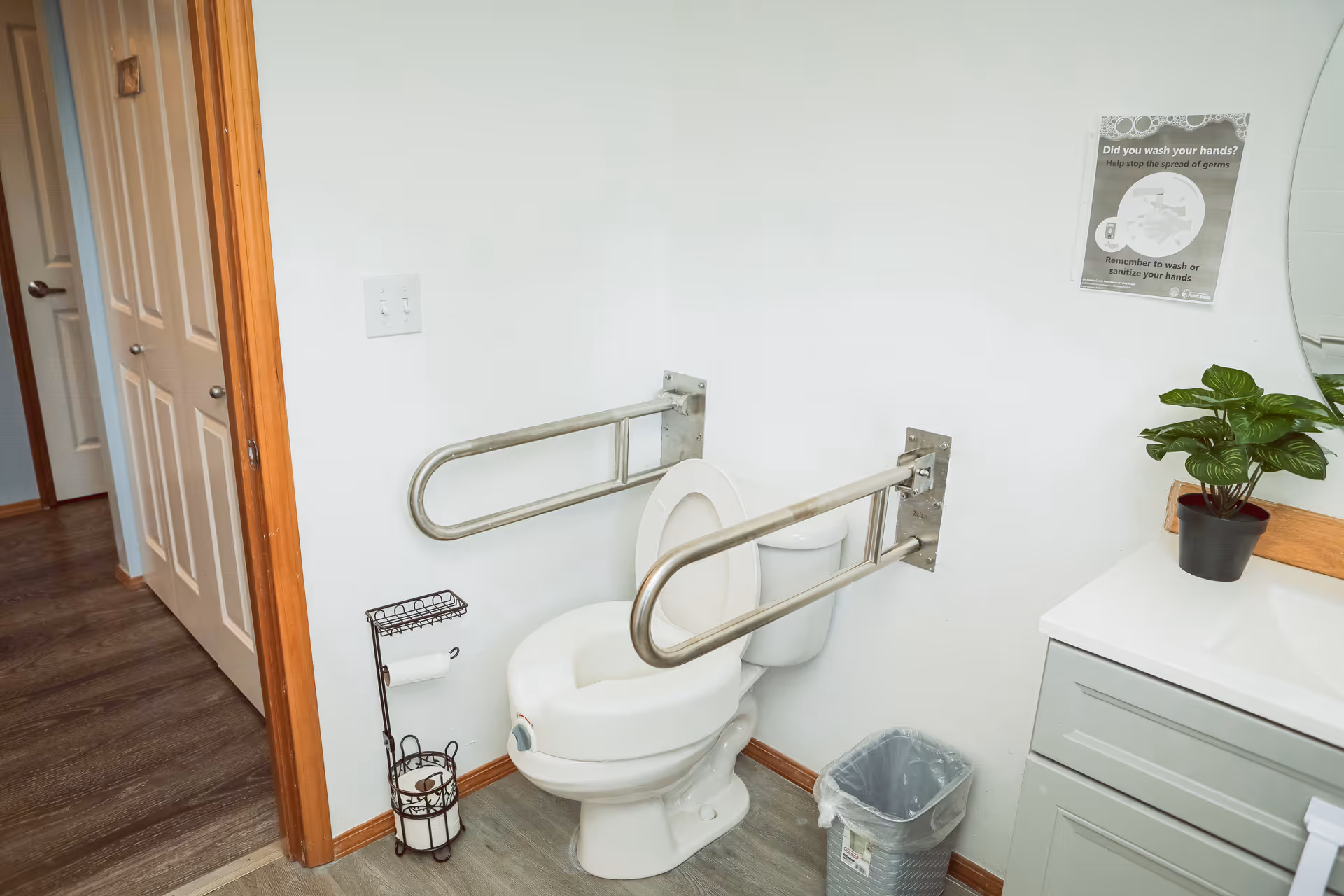 A bathroom with a white toilet equipped with metal grab bars on both sides for accessibility. There is a toilet paper holder with extra rolls next to the toilet, a small trash can, a sink with a cabinet, a potted plant on the counter, and a round mirror on the wall. A sign reminding to wash hands is also visible on the wall.