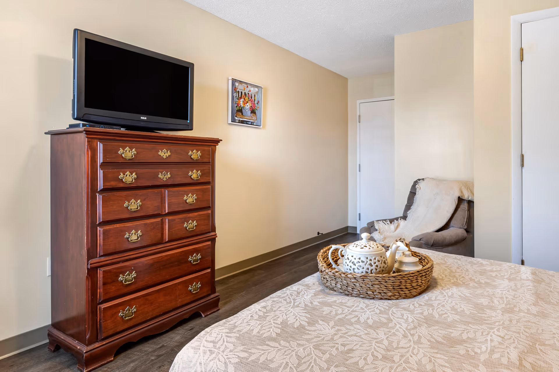 A bedroom with a wooden dresser topped with a flat-screen TV, a beige wall with a small framed painting, a cozy armchair with a throw blanket, and a bed with a decorative tray holding a teapot and cups.