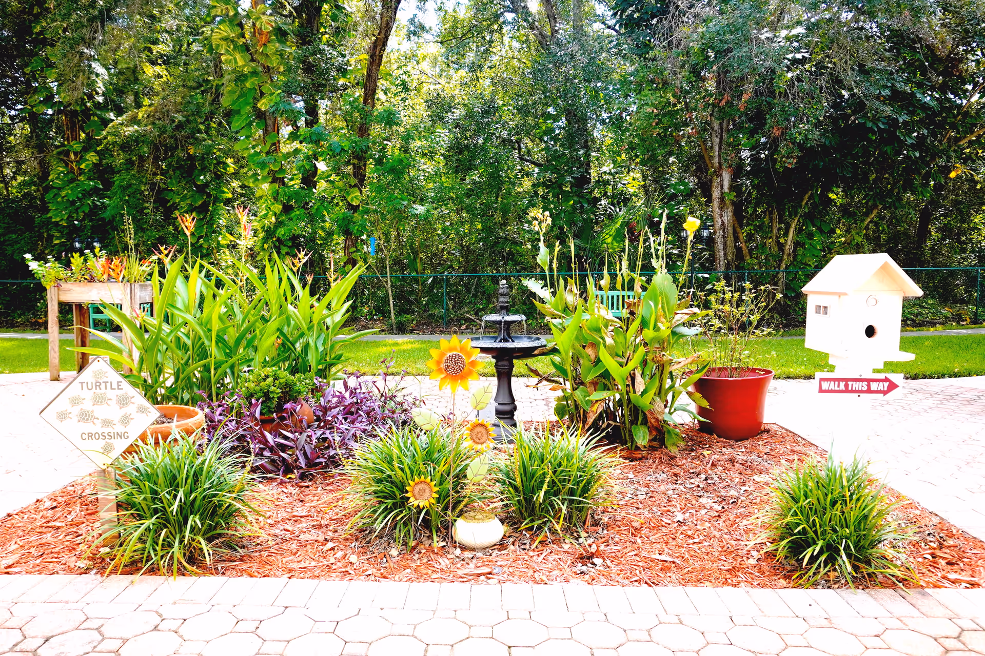 A small garden area with various green plants and flowers, including decorative sunflower ornaments. There is a black birdbath in the center, a wooden birdhouse on a post with a 'WALK THIS WAY' sign, and a 'TURTLE CROSSING' sign. The garden is surrounded by a paved walkway and dense trees in the background.