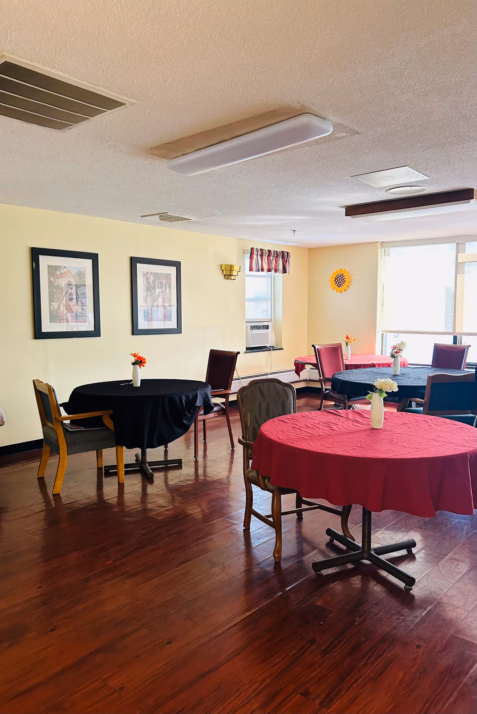 A dining room with several round tables covered with red and black tablecloths, each table having a small vase with flowers. The room has wooden floors, light yellow walls with two framed pictures, a window with an air conditioning unit, and a large window letting in natural light.
