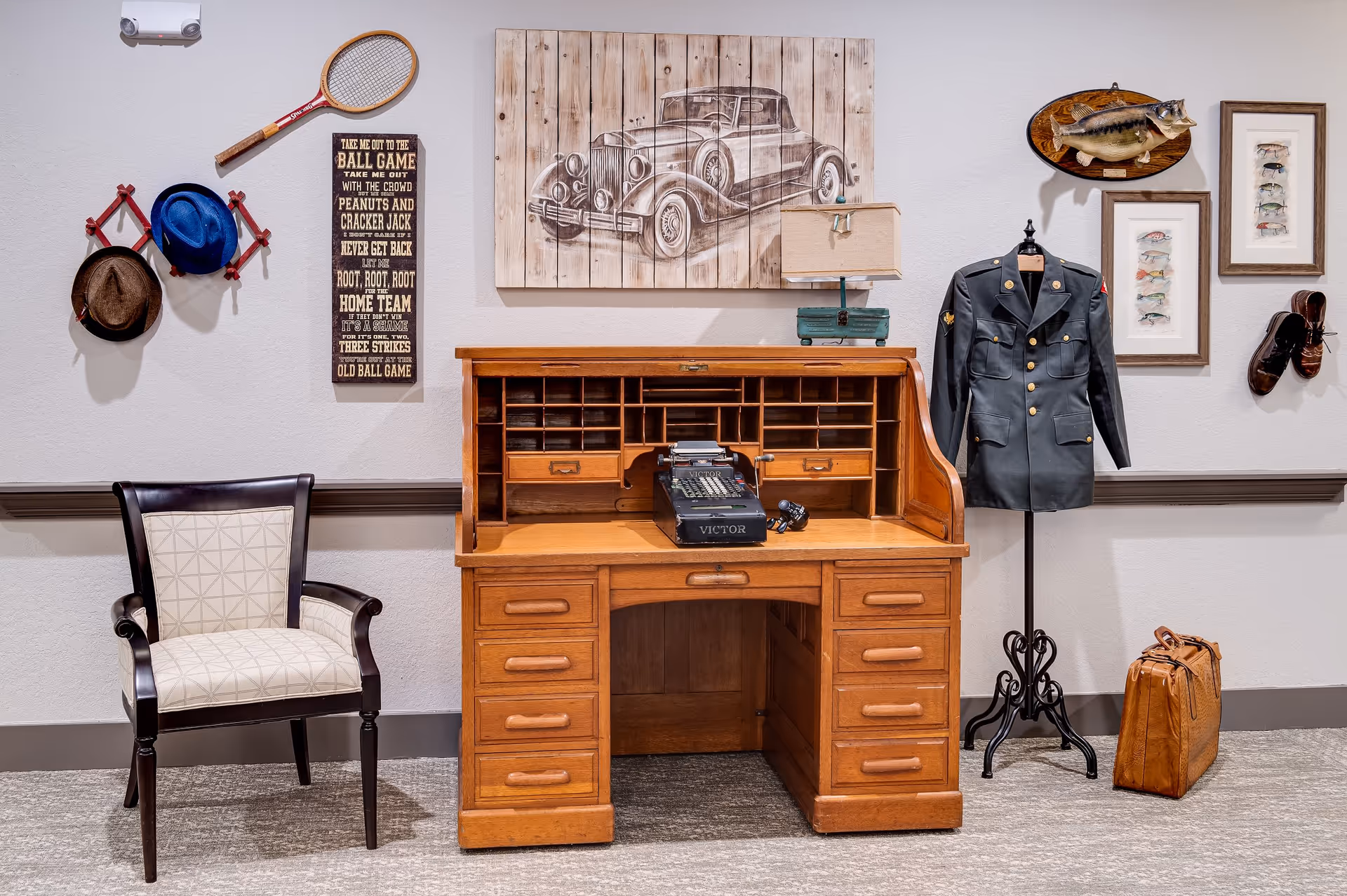 A vintage-themed interior corner featuring a wooden roll-top desk with an old typewriter and a small lamp on top. To the left of the desk is a wooden chair with a cushioned seat and backrest. On the wall above the chair, there are two hats hanging on a red rack and a wooden tennis racket. A vertical sign with baseball-related text is also mounted on the wall. To the right of the desk, a mannequin dressed in a military uniform stands next to a brown leather bag on the floor. The wall behind has framed pictures of fish and a mounted fish plaque, along with a pair of brown shoes hanging on the wall.