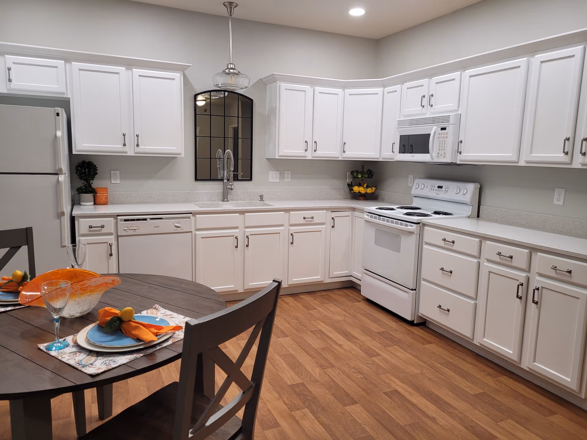 A modern kitchen with white cabinets and appliances including a refrigerator, dishwasher, stove, and microwave. There is a round wooden dining table with two place settings, orange napkins, and a decorative orange glass centerpiece. The floor is wood, and the walls are light-colored with a window-like mirror above the sink.