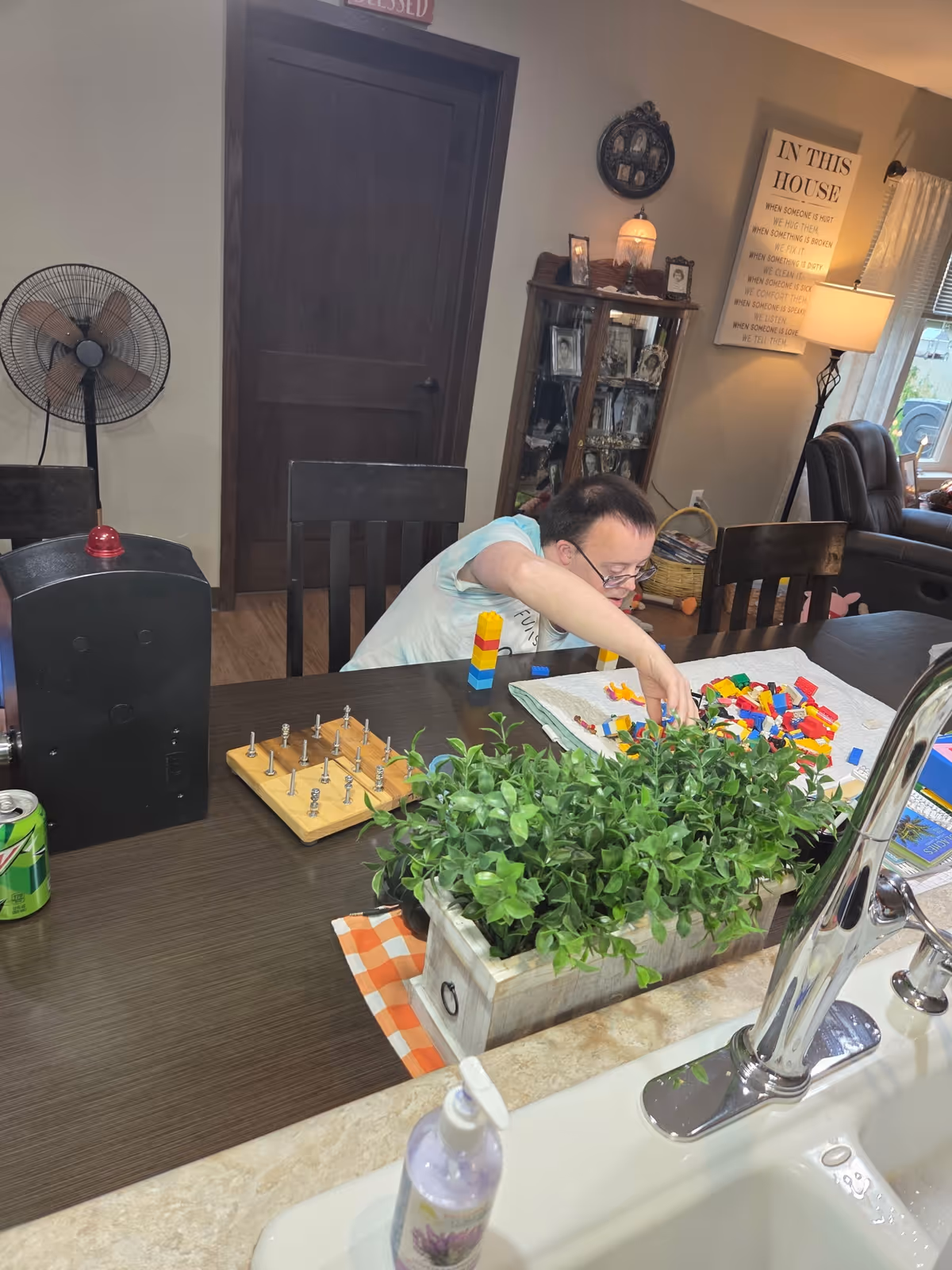 A person sitting at a dining table playing with colorful building blocks. The table has a wooden game with pegs, a can of soda, and a rectangular planter with green plants. In the foreground, there is a kitchen sink with a soap dispenser. The background shows a closed wooden door, a standing fan, a glass cabinet with framed photos, a lamp, and a wall sign with text.