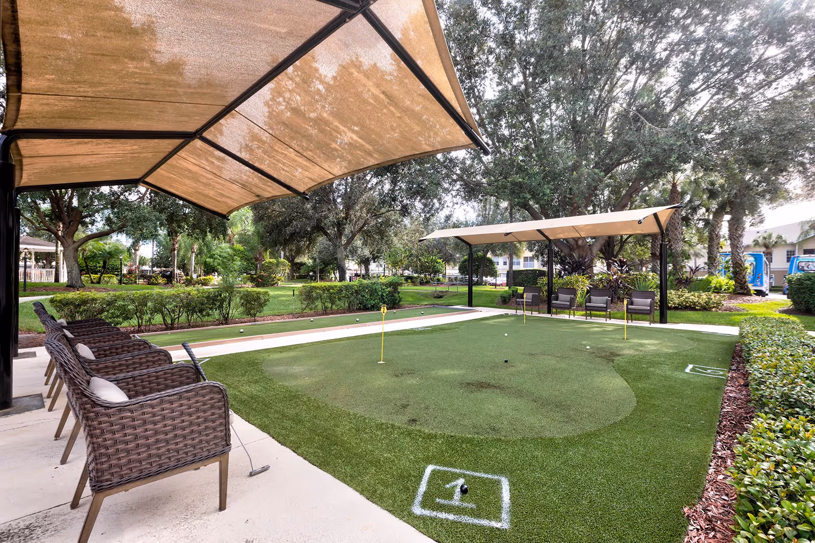 Outdoor putting green area with several wicker chairs under a large canopy on the left and additional chairs under another canopy on the right, surrounded by trees and shrubs in a garden setting.