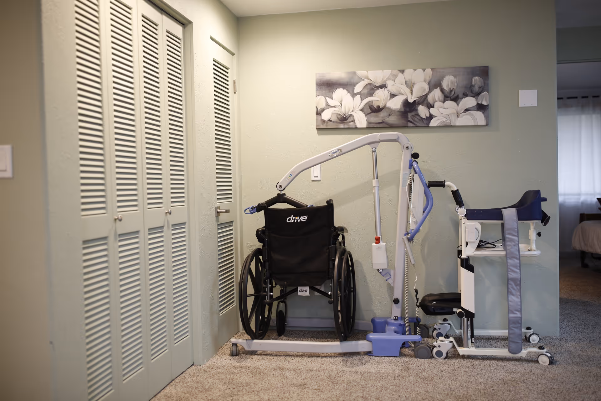 A corner of a room with a wheelchair, a patient lift, and a standing aid device placed against a light green wall. Above them is a rectangular floral painting with white flowers. To the left are two louvered closet doors and a regular door. The floor is carpeted, and part of a bed and window with curtains are visible in an adjacent room.