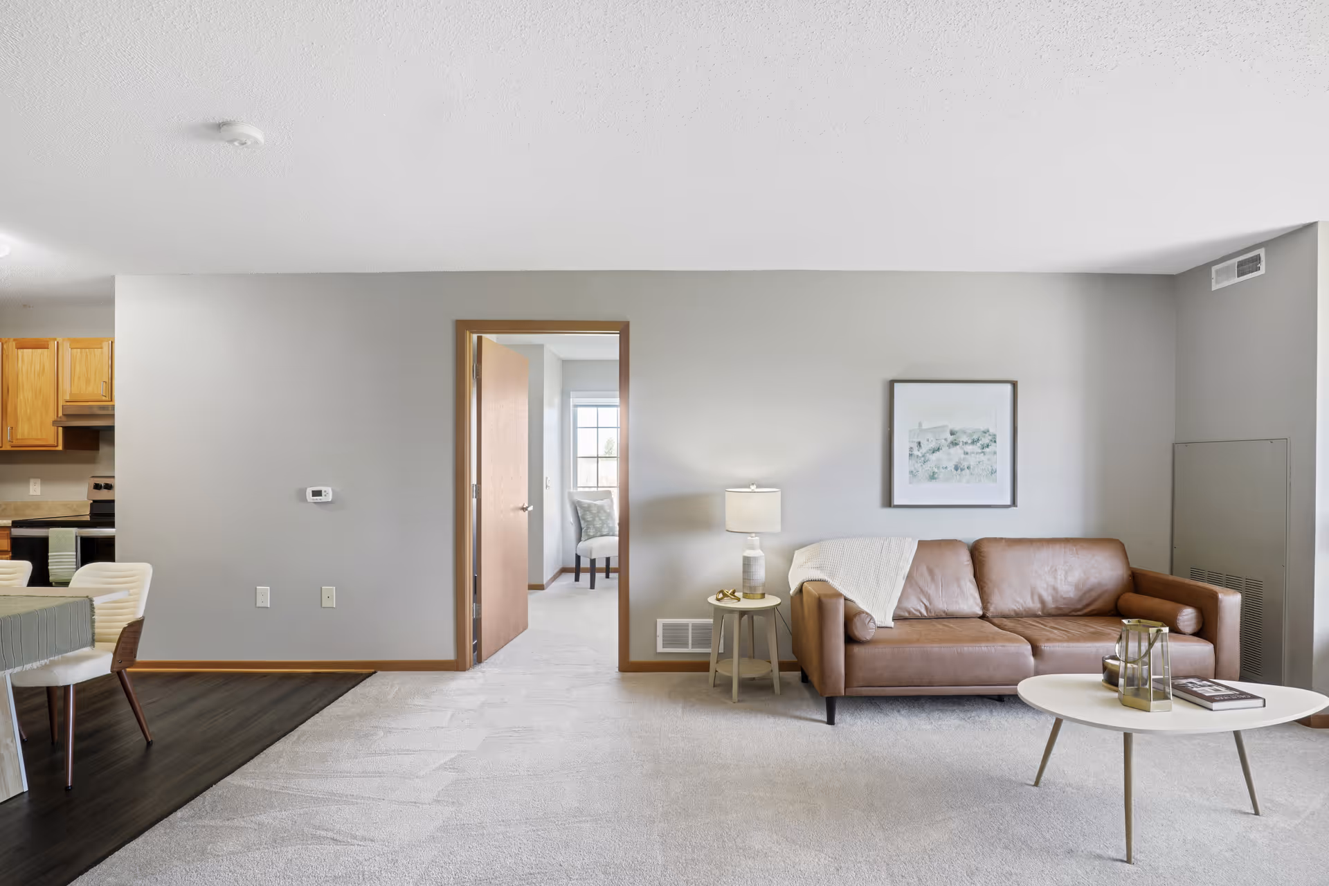 A bright living room with a brown leather sofa, a small round white coffee table with decorative items, and a side table with a lamp. The walls are painted light gray, and there is a framed picture hanging above the sofa. To the left, there is an open doorway leading to another room with a chair by a window. Part of a kitchen and dining area with wooden cabinets and white chairs is visible on the far left.