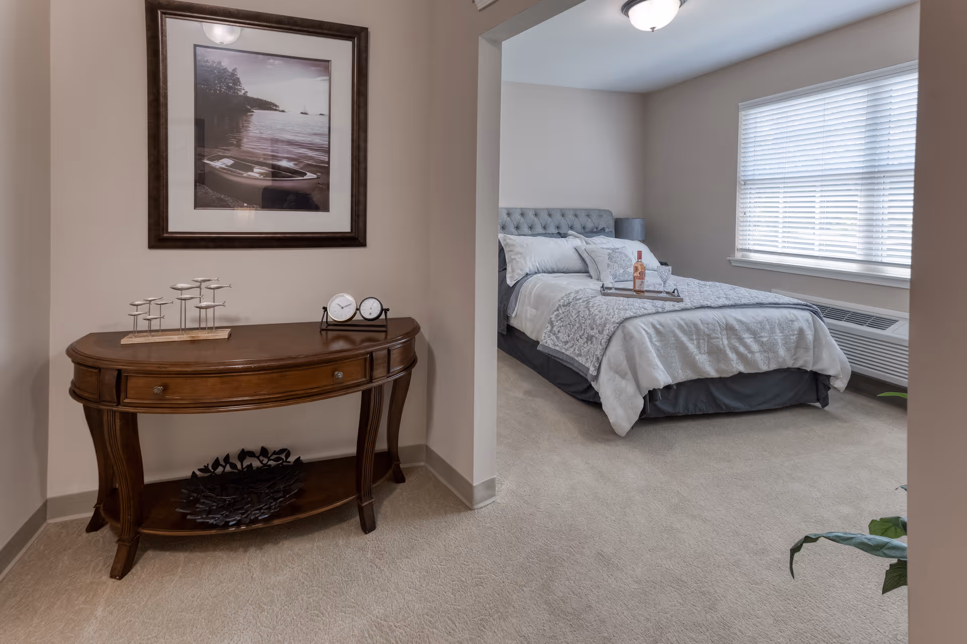 A cozy bedroom with a neatly made bed featuring white and gray bedding, a tray with a bottle and glass on the bed, a large window with blinds, and a wooden console table with decorative items and a framed picture of a canoe on a lake hanging above it.