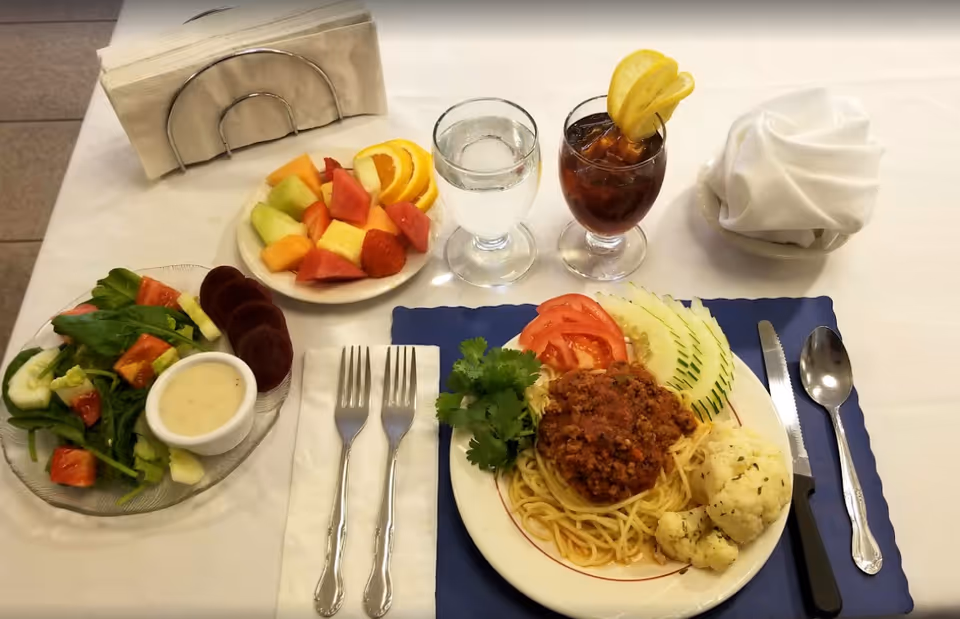 A neatly arranged meal on a white tablecloth featuring a plate of spaghetti with meat sauce, sliced tomatoes, cucumber, and cauliflower, a side salad with dressing, a plate of mixed fresh fruit, a glass of water, and a glass of iced tea garnished with lemon slices. Two forks, a knife, and a spoon are placed on either side of the main plate, with a napkin holder and a folded white cloth napkin also visible.