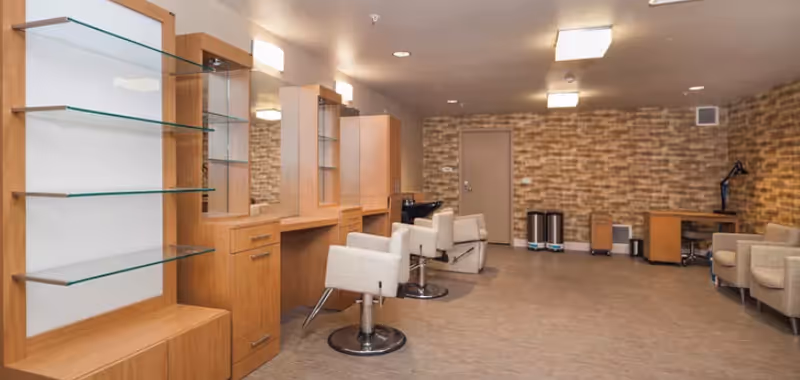 Interior view of a salon area in a senior living facility with wooden styling stations, mirrors, white cushioned salon chairs, and a brick accent wall. There are also two beige armchairs and a small desk with a lamp in the corner.