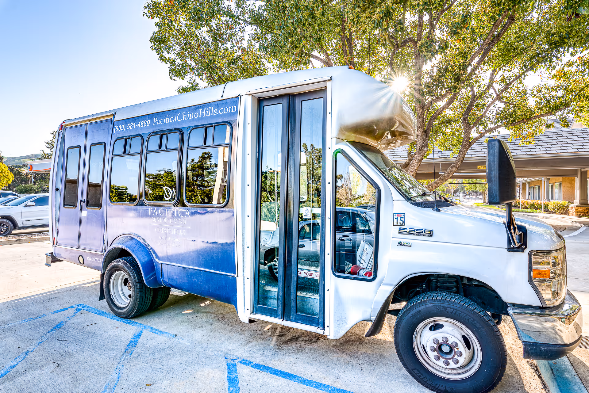 A white and blue shuttle bus parked in a parking lot near a senior living facility. The bus has the text 'Pacifica Chino Hills' and a phone number on the side. Trees and part of the building are visible in the background.