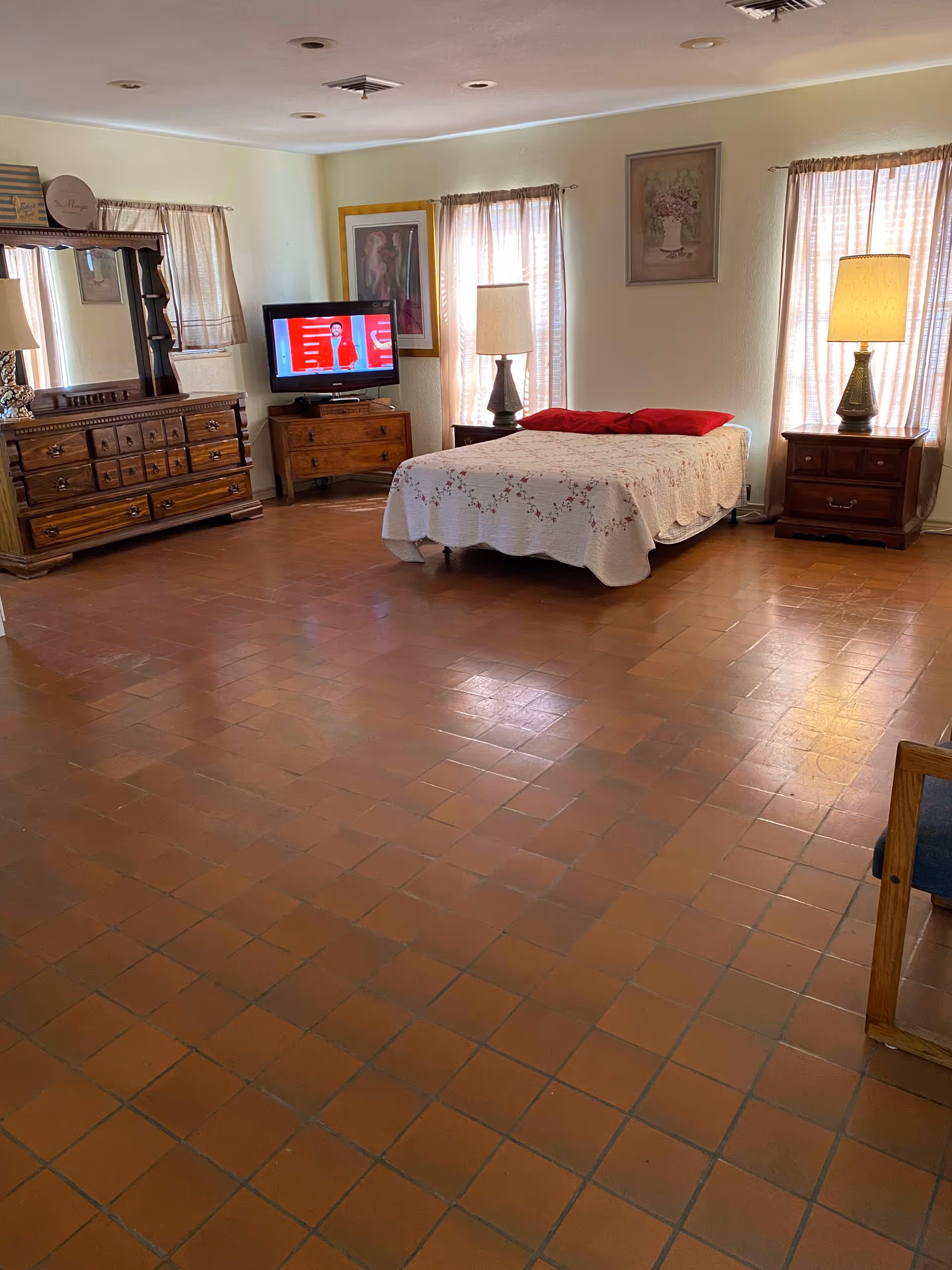 A spacious bedroom with terracotta tiled floor, a bed with a white floral quilt and red pillows, two wooden nightstands each with a lamp, a wooden dresser with a large mirror, a small TV on a wooden stand, and three windows with sheer curtains letting in natural light. The walls are light-colored and decorated with framed artwork.