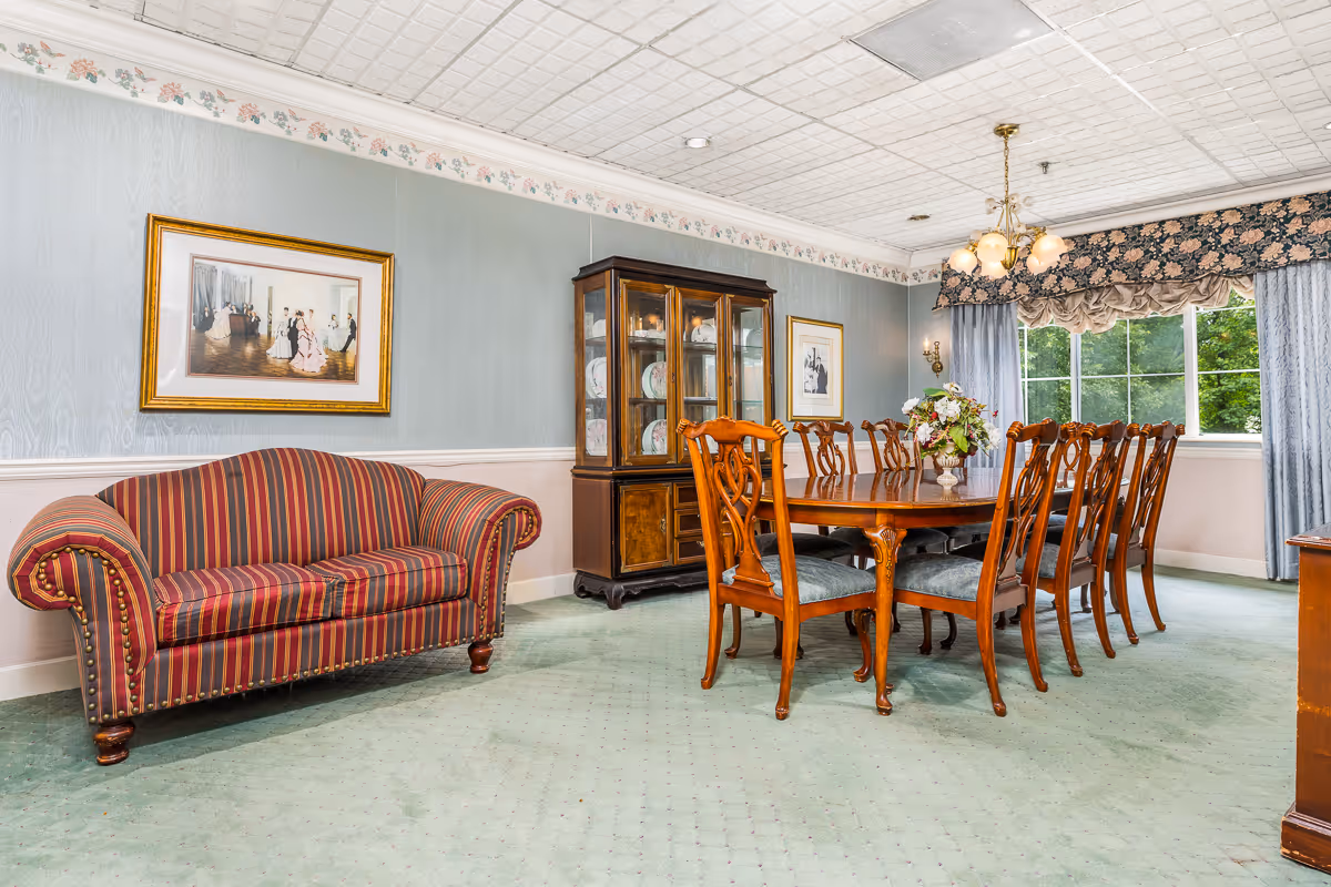A formal dining room with a long wooden table and chairs, a china cabinet, and a striped sofa beneath framed artwork.