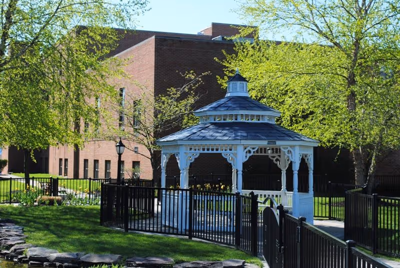 A white gazebo with a blue-gray roof is situated in a fenced garden area with green grass and trees. Behind the gazebo, there is a large brick building with multiple windows. The scene is bright and sunny with clear skies.