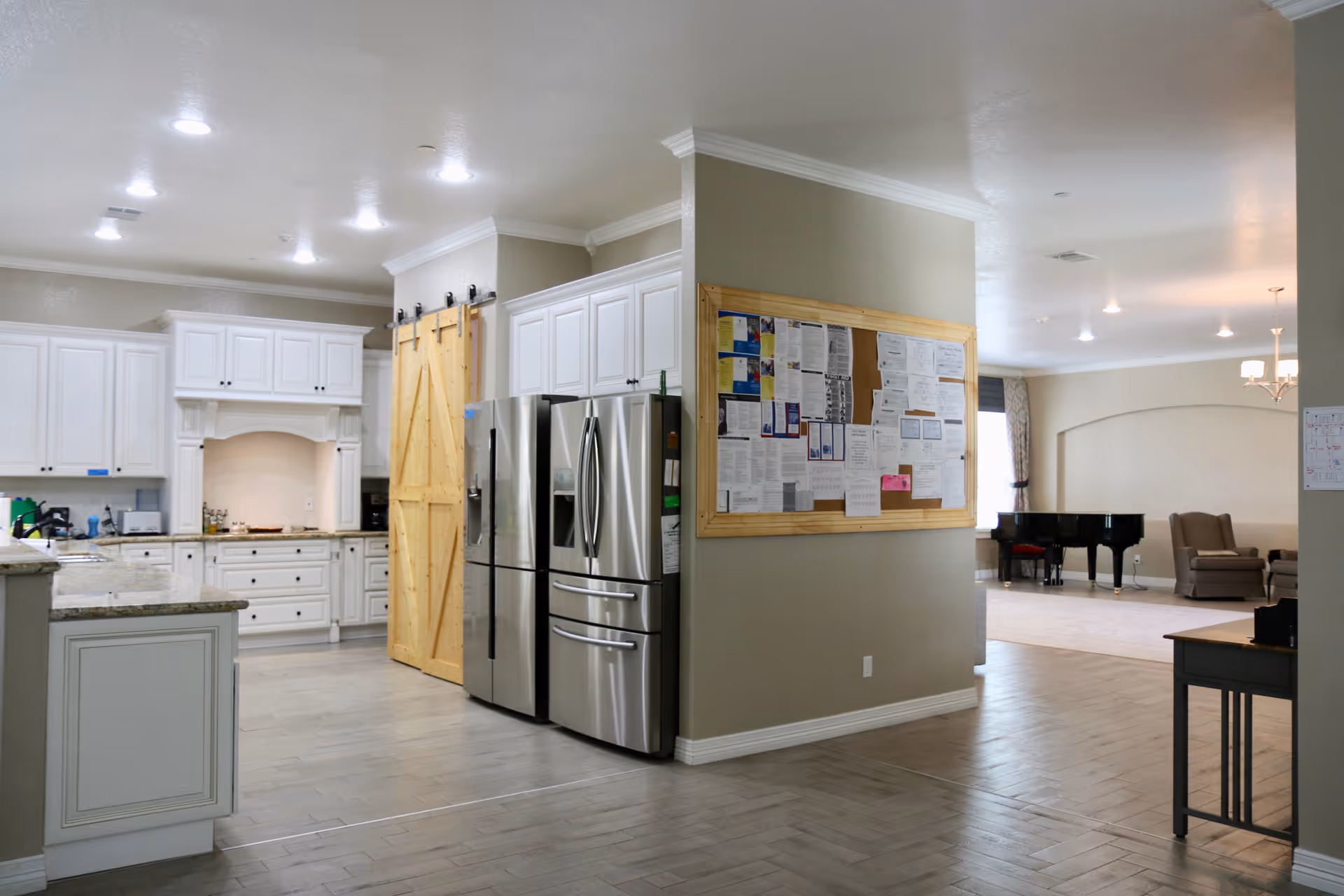 Interior view of a senior living facility showing a modern kitchen area with white cabinets, granite countertops, and two stainless steel refrigerators. A wooden sliding barn door is visible next to the refrigerators. Adjacent to the kitchen is a bulletin board filled with various notices and papers. In the background, there is a living area with a grand piano, armchairs, and a chandelier, all set on light-colored flooring under recessed ceiling lights.
