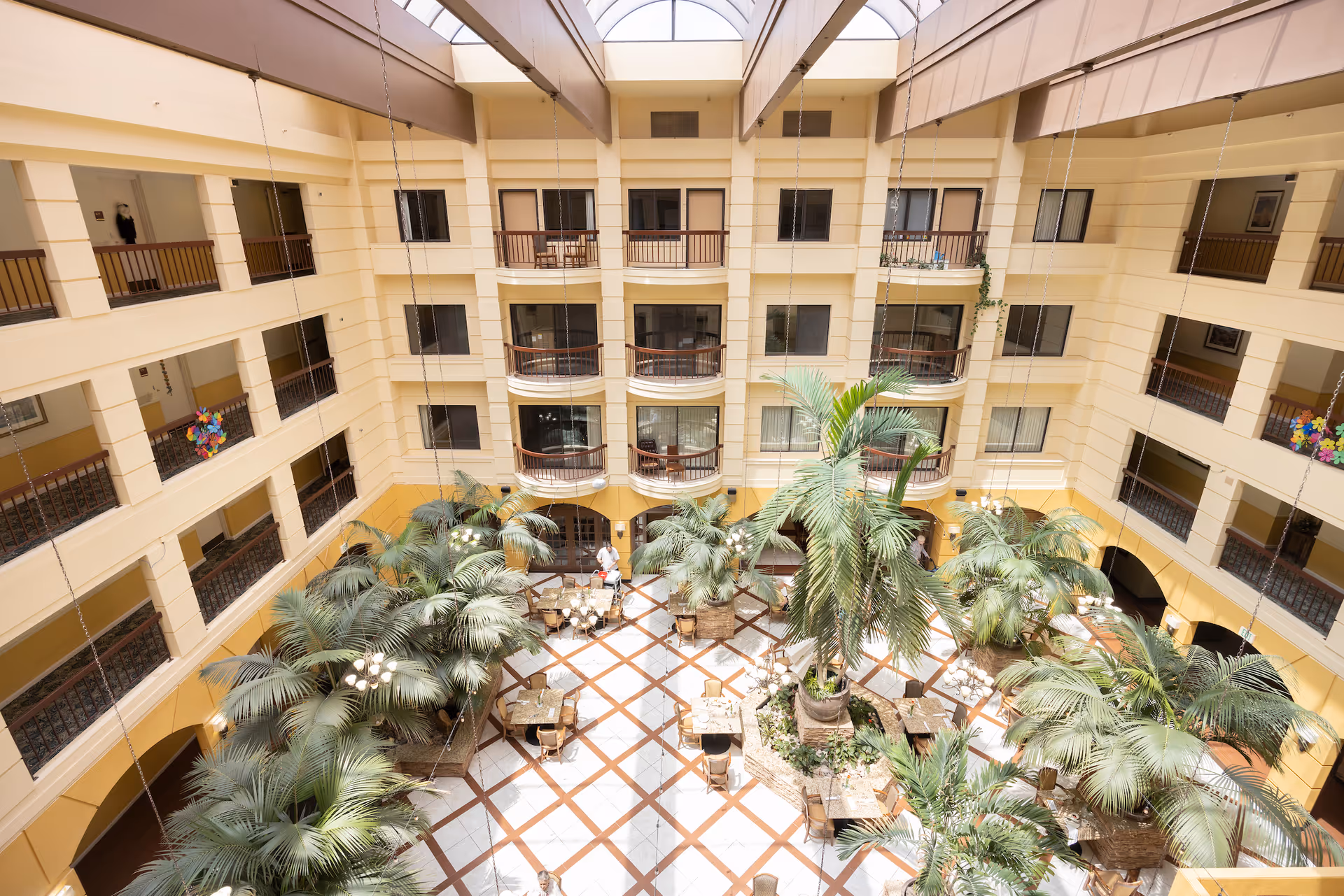 Interior view of a senior living facility atrium with multiple floors of rooms and balconies surrounding a central open area filled with large potted palm plants and several tables and chairs. The space is well-lit with natural light coming from a skylight above.