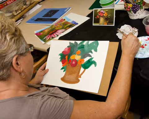 An elderly woman with short blonde hair and glasses is painting a colorful still life of a brown vase with flowers and fruit on a white sheet of paper. She is seated at a table with art supplies, including paintbrushes, a palette, and reference photos of flowers.