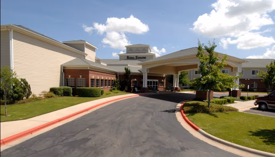Exterior view of a senior living facility with a covered entrance driveway, manicured lawns, bushes, and trees under a partly cloudy sky.