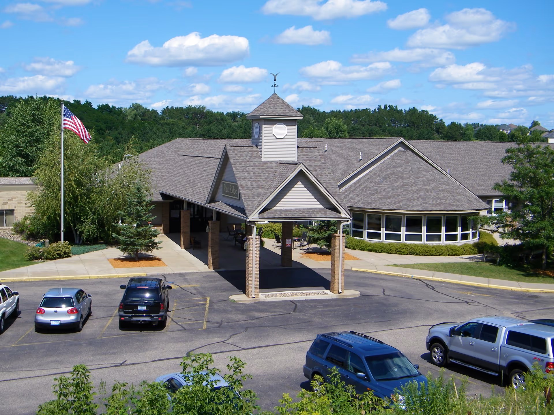 Entrance of a single-story senior living building with a covered porte-cochere, American flag, and parked cars in the lot.
