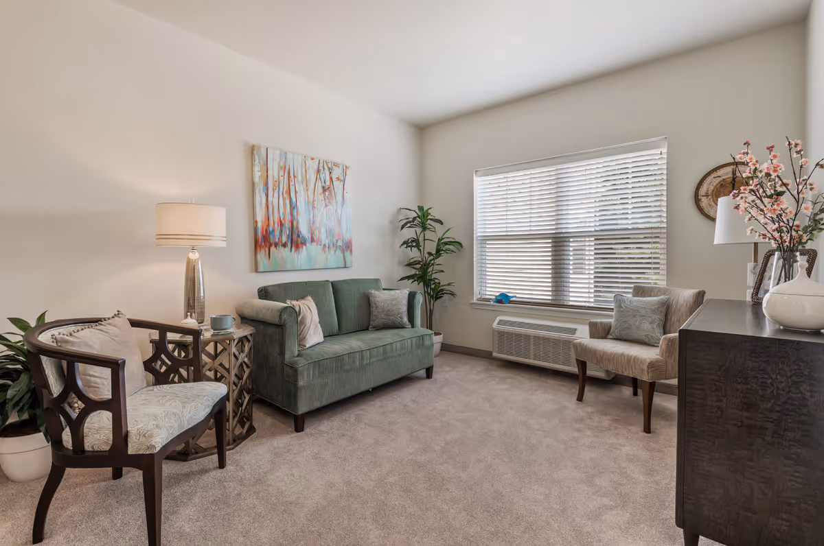 A cozy living room with a green upholstered sofa, two patterned armchairs, a side table with a lamp and a cup, a large window with blinds, a potted plant, a wooden cabinet with a white vase holding pink flowers, and a colorful abstract painting on the wall.