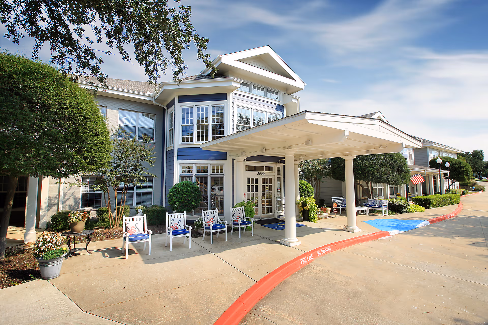 Senior living facility entrance with a covered drop-off, chairs, and manicured landscaping.