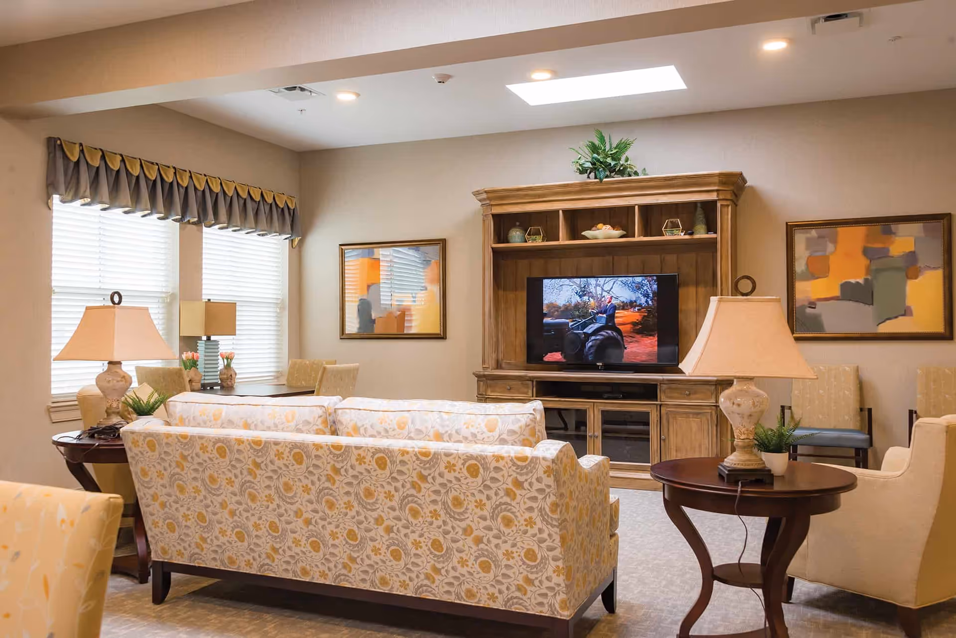 A cozy living room with a floral patterned sofa facing a wooden entertainment center with a TV. The room has beige walls, two framed abstract paintings, two table lamps on round wooden side tables, and a window with blinds and a valance.