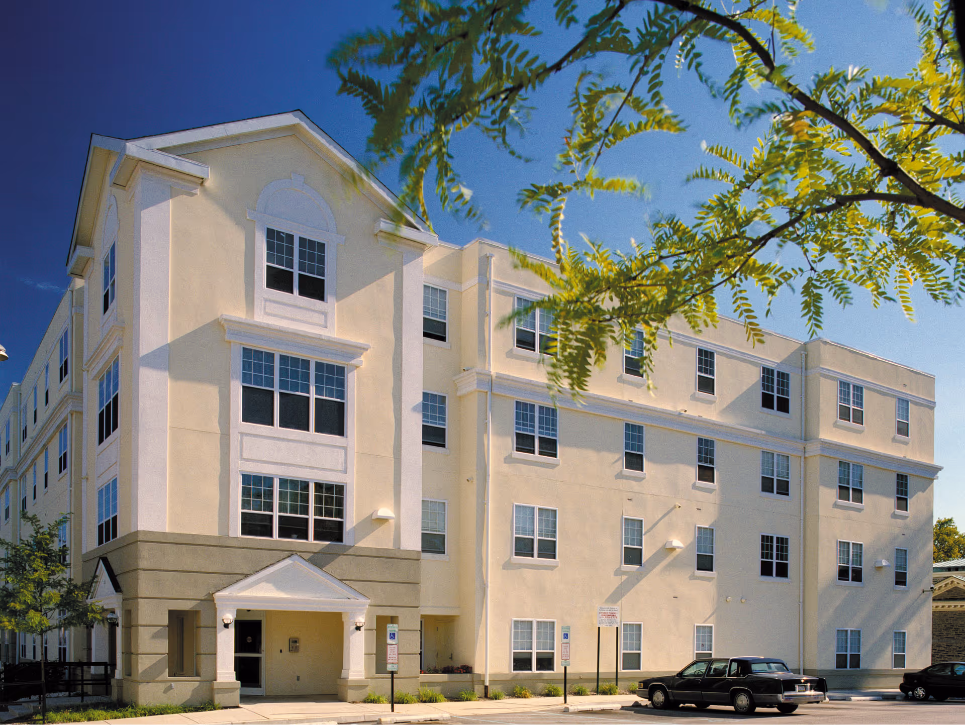 Exterior view of a four-story beige and white senior living facility building named Maple Village, with multiple windows, a small entrance with a white triangular roof, a few parked cars, and green tree branches in the foreground under a clear blue sky.