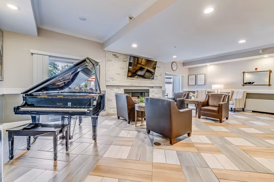 A bright and spacious senior living common area featuring a black grand piano with a matching bench on the left, several brown leather armchairs arranged around wooden tables, a stone fireplace with a mounted flat-screen TV above it, and large windows letting in natural light. The room has a patterned tile floor and neutral-colored walls with framed artwork and a clock.