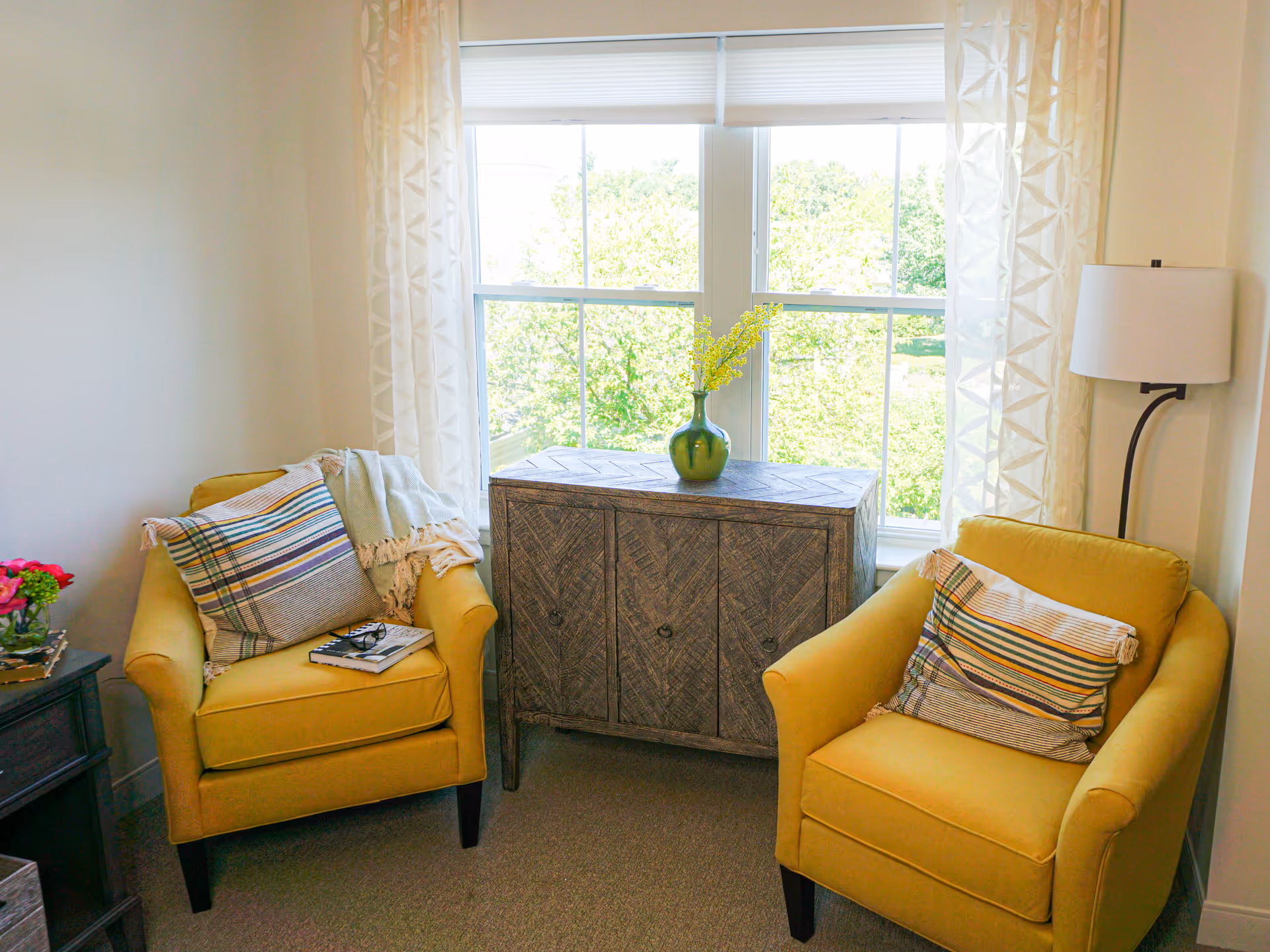 Two yellow armchairs with striped pillows flank a wooden cabinet beneath a bright window in a cozy sitting area.