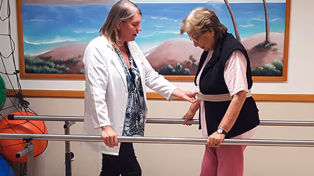 A healthcare professional assists an elderly woman with walking exercises using parallel bars in a rehabilitation or therapy room. A colorful mural of a beach scene is visible on the wall behind them.