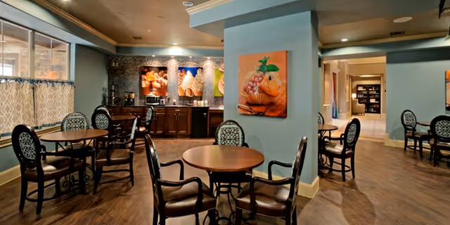 Dining/lounge area with round wooden tables and chairs, a small kitchenette along the back wall, and colorful artwork on teal walls.