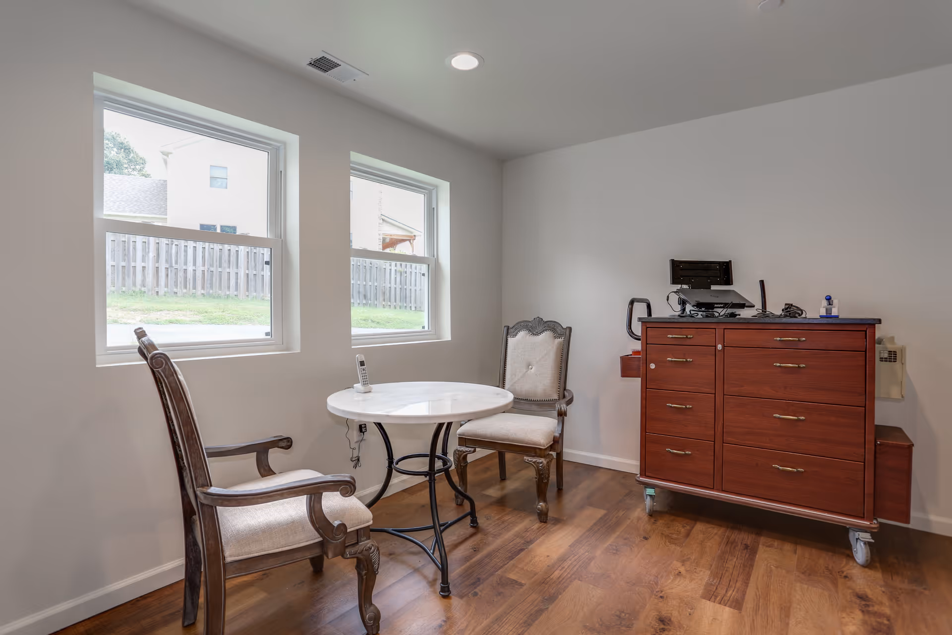 Small sitting room with two upholstered chairs around a round table by two windows and a wooden dresser on casters.