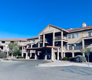 Exterior view of a multi-story senior living facility building with a covered entrance, balconies, and surrounding landscaping under a clear blue sky.
