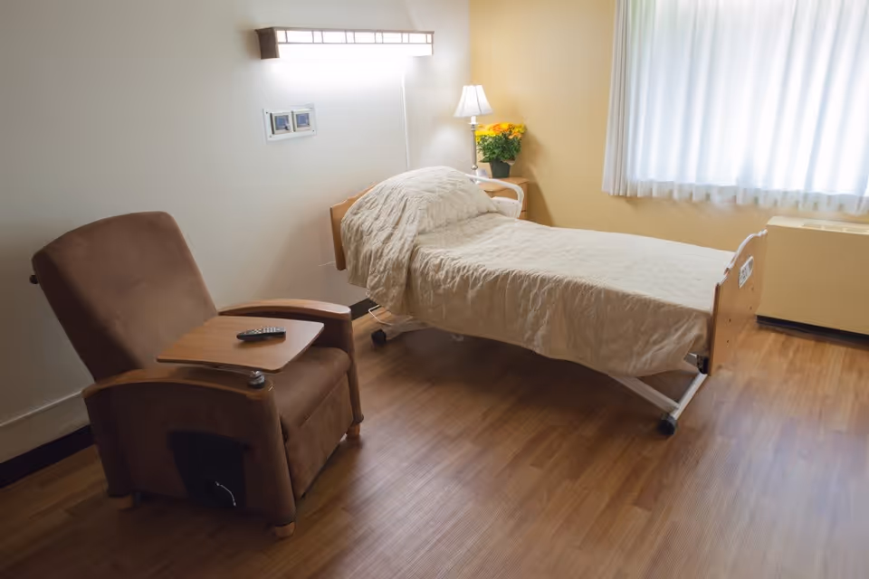 A simple, clean bedroom in a senior living facility with a single bed covered in a beige quilt, a brown recliner chair with a wooden tray and remote control, a small bedside table with a lamp and a pot of yellow flowers, and a window with white curtains letting in natural light.