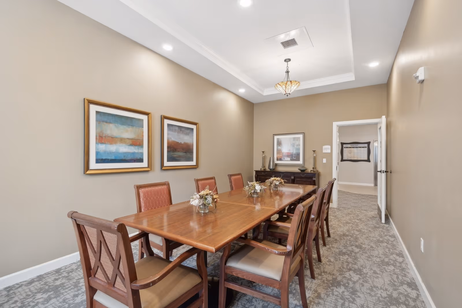 A formal dining room with a long wooden table surrounded by eight wooden chairs with cushioned seats. The walls are beige with two framed abstract paintings on the left wall and another framed picture on the far wall above a dark wooden sideboard. The room has recessed lighting and a decorative ceiling light fixture. The floor is carpeted with a patterned design, and an open doorway leads to another room.