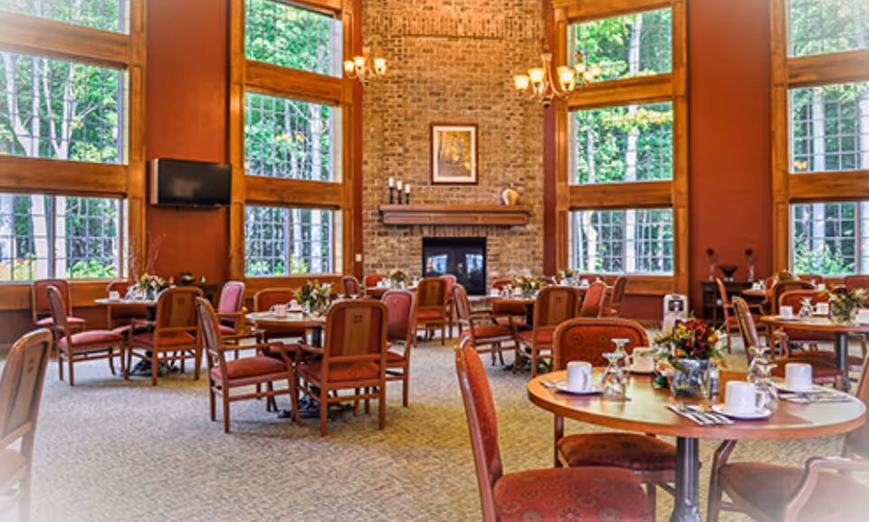 A spacious dining room with multiple round tables set with white cups, glasses, and silverware. The room features large windows with wooden frames letting in natural light and views of green trees outside. There is a brick fireplace with a mantel and a framed picture above it. The walls are painted in warm tones, and the chairs have red upholstery with a subtle pattern.