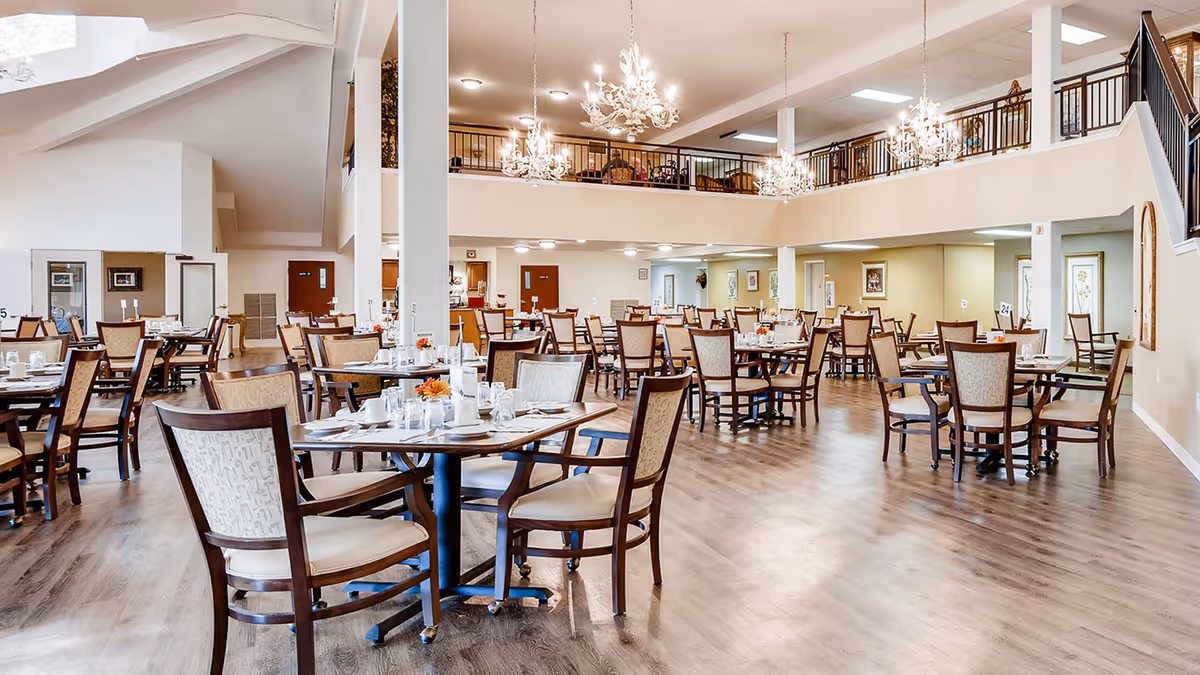 Spacious dining room in a senior living community with multiple round tables and chairs arranged neatly. The room features high ceilings, chandeliers, and a second-floor balcony overlooking the dining area. Tables are set with white tableware and small floral centerpieces.