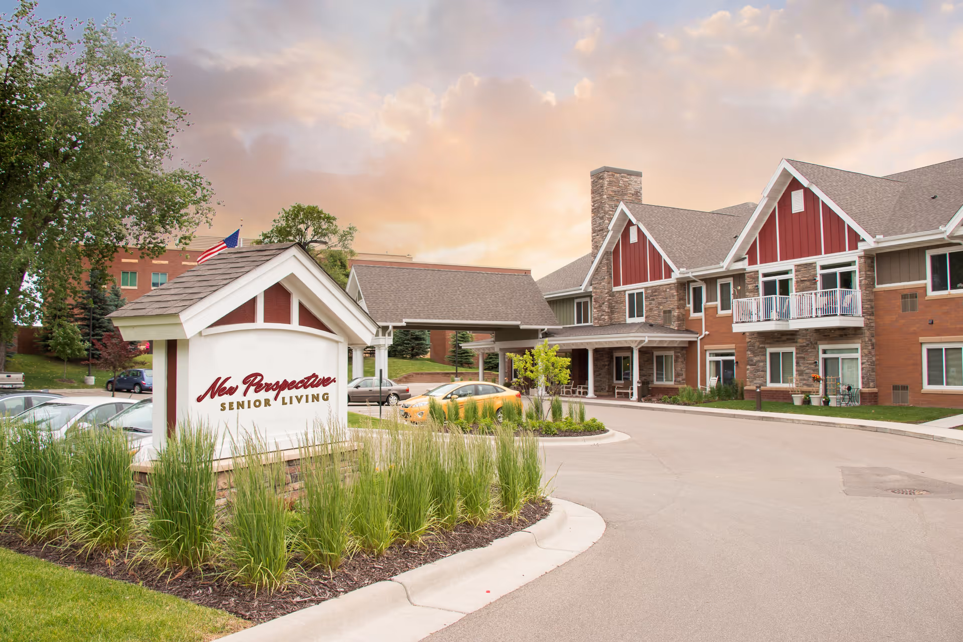 Exterior view of New Perspective Senior Living facility in Woodbury during sunset, showing a large building with red and beige siding, multiple windows, a covered entrance, landscaped greenery, and a sign with the facility name near the driveway.