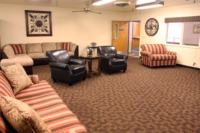 A spacious living room area with a brown patterned carpet, featuring a beige sectional sofa, two black leather armchairs around a small round wooden table with a decorative vase, and two striped armchairs. The walls are light-colored with framed artwork, a window with blinds, and a door leading to another room.