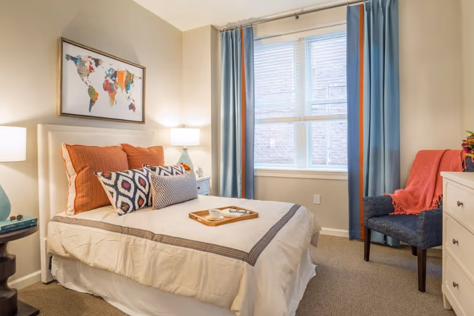 A cozy bedroom with a neatly made bed featuring beige bedding and multiple decorative pillows in orange and patterned designs. A wooden tray with a cup and glasses rests on the bed. The room has a window with blue and orange curtains, a blue armchair with an orange throw blanket, a white dresser, and two bedside tables with lamps. A colorful world map artwork hangs above the bed.
