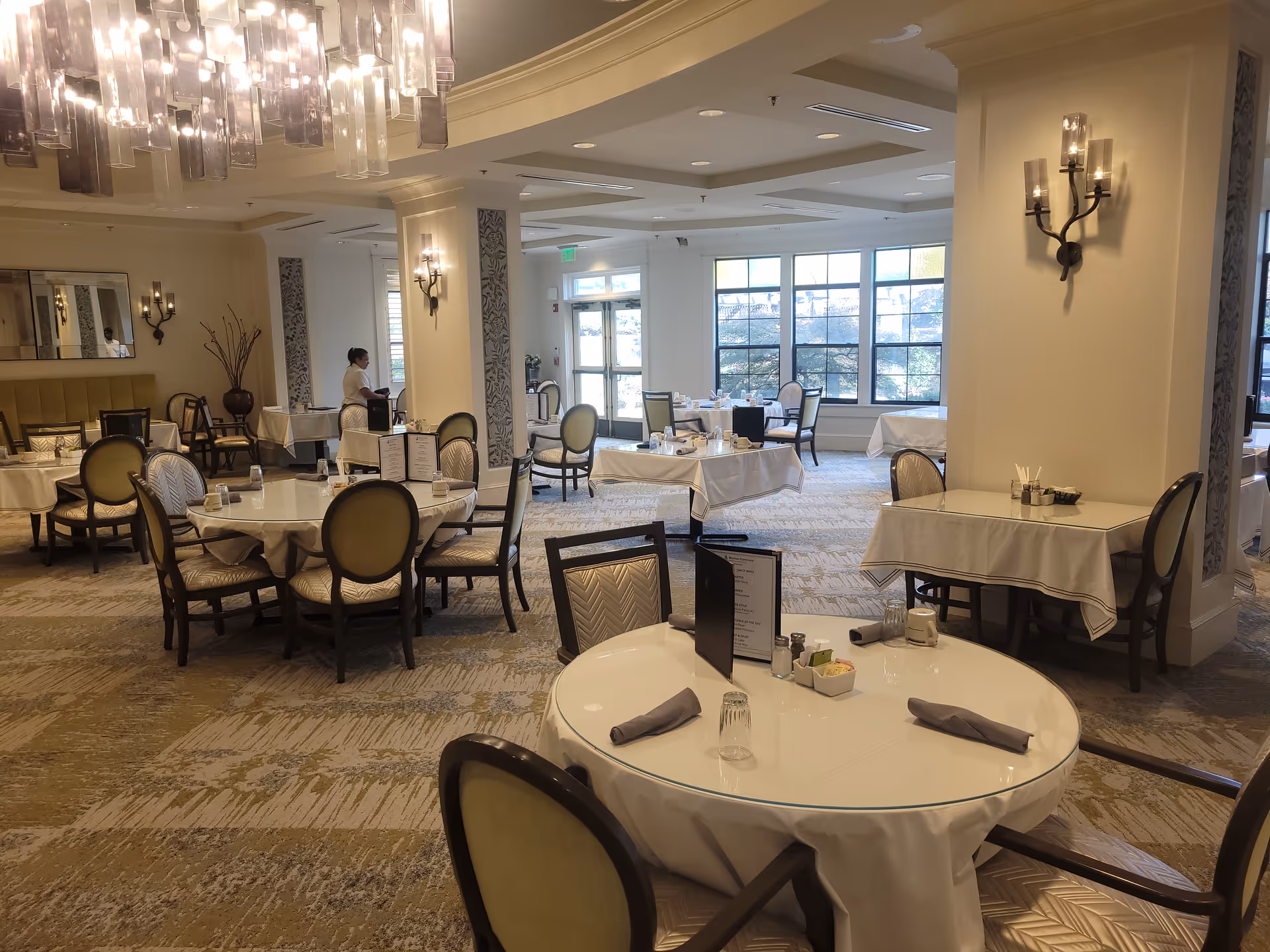 Spacious, well-lit dining room with round tables set with white tablecloths and upholstered chairs.