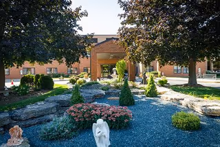 Outdoor garden area with a gravel path, small shrubs, flowers, and a white angel statue in the foreground. Large trees provide shade, and a brick building with an entrance canopy is visible in the background.