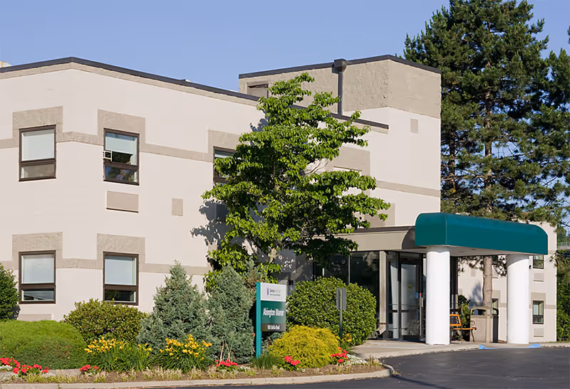 Exterior view of a two-story beige building with a green awning over the entrance. The building is surrounded by well-maintained landscaping including bushes, trees, and flowers. A sign near the entrance reads 'Abington Manor'.